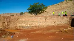 Grande área escavada no meio da montanha. Ao longe, é possível ver várias pessoas usando coletes amarelos.