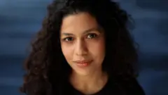 Arianna de Souza-García mira a la cámara. Es una mujer de cabello rizado y oscuro, con camiseta negra, situada frente a un fondo azul oscuro.