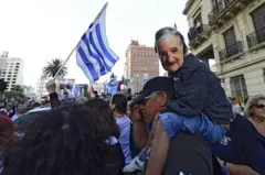 Multidão para receber Mujica, com a bandeira do Uruguai, e uma com uma máscara do ex-presidente uruguaio nas costas de um adulto