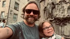 La imagen muestra a Darren y Sophia Scott en una selfie. Él tiene el cabello rubio, algo largo, y barba; lleva gafas de sol y una camiseta gris. Tiene el brazo alrededor de Sophia, quien lleva gafas y una bufanda. Ambos están sonriendo.
Detrás de ellos se aprecia un monumento o escultura de piedra desgastada, con figuras talladas y lo que parece ser una cresta o un escudo de armas.