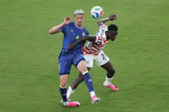 Dois jogadores de futebol, um de camisa azul e Folarin Balogun de vermelho e branco, disputa a bola, quando Balogun agarra a camisa do outro jogador