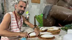 Cristiano Bara olhando para a câmera e sorrindo, enquanto manipula adereços na mesa