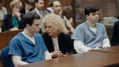 Di Menendez brothers, wearing different shades of a blue prison uniform, sit at a table wit one of dia attorneys during dia trial for 1994. Inside di courtroom, dia lawyer na woman wit curly hair and appear deep in conversation wit Erik Menendez. 