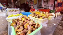 Puesto de mercado exhibe bandejas llenas de hongos y verduras frescas en una calle comercial.