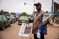 Un homme tenant des journaux dans ses mains debout au bord d'une route avec des voitures à l'arrière-plan