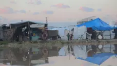 ガザの難民キャンプの仮設テントの前が、広範囲に冠水している。テントの間に子ども2人が立っている。テントの柱には細い木材が、屋根の代わりにビニールシートが使われている。テントの側面の一部は布やビニールシートで覆われているが、片側には壁がなく、外からテント内部が見えている