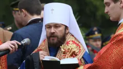 Мужчина в митрополичьем клобуке читает книгу в микрофон