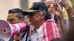 Gustavo Petro com um megafone na mão em uma assembleia pró-palestina em Nova York. Ele está de bone azul, óculos escuros, camisa branca e um tecido bordado vermelho e branco nas costas