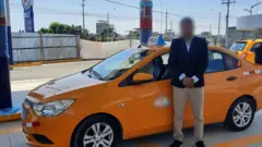 Um homem de blazer azul marinho e calça bege, com o rosto borrado, aparece diante de um táxi em um posto de gasolina.