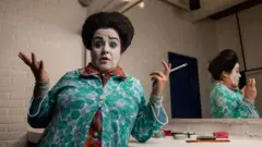 Gabriela Muñoz en un camerino, con un pincel en una mano, muestra a la cámara su maquillaje del personaje Chula: con una ceja levantada, un corazoncito pintado en los labio y un grueso pelo crespo. Un espejo la refleja de ángulo.