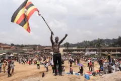 Kijana mtanashati anasimama juu ya kilima kidogo cha uchafu, akipeperusha bendera ya Uganda. Nyuma yake ni umati wa watu katika mkutano ulioongozwa na kiongozi wa upinzani, Robert Kyagulanyi Ssentamu.
