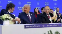 Дональд Трамп, який заснував нову міжнародну організацію, сидить за столом і тисне руки іншим учасникам зібрання. 