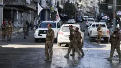 Suriye'nin askeri polis mensupları bir sokakta yürüyüş yapıyor. Arka planda araçlar var. 