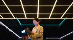 Una mujer con gafas transparentes y una tableta en la mano, en un salón oscuro con varias lámparas de luz fluorescente en el techo, las cuales le dan una sensación futurista a la foto.