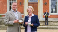 Casal rico toma café em frente a mansão. Por trás, um mordomo