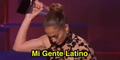 A cantora Jennifer Lopez segurando um prêmio acima da cabeça, com a cabeça levemente abaixada e expressão emocionada. Ela usa um vestido preto com detalhes rendados. Sobre a imagem aparece o texto: “Mi Gente Latino”.