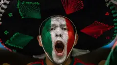 Un hombre con la boca abierta, seguramente está gritando o cantando. Tiene la cara pintada con los colores verde, blanco y rojo, que conforman la bandera de México. Lleva puesto un sombrero de mariachi.