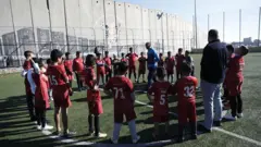 Um grupo de crianças vestindo uniforme de futebol está parado no meio do campo, em círculo, enquanto ouve o treinador falar