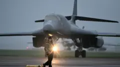 7 मार्च 2026 को सूर्योदय के तुरंत बाद दक्षिण-पश्चिम इंग्लैंड के RAF फ़ेयरफ़ोर्ड एयरबेस पर अमेरिकी वायुसेना के B-1 लांसर बॉम्बर के सामने एक ग्राउंड क्रू सदस्य खड़ा है. ब्रिटेन के प्रधानमंत्री किएर स्टार्मर ने वॉशिंगटन को हिंद महासागर में स्थित डिएगो गार्सिया और दक्षिण-पश्चिम इंग्लैंड के RAF फ़ेयरफ़ोर्ड एयरबेस का इस्तेमाल ईरान के मिसाइल ठिकानों पर बमबारी करने के लिए करने की मंज़ूरी दे दी है. यह फैसला तब लिया गया जब ईरान की जवाबी कार्रवाइयों में खाड़ी के कई देशों को निशाना बनाया गया.