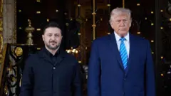 Зеленський і Трамп стоять поруч на сходах перед великою декоративною металевою брамою з позолоченими елементами. Ліворуч — людина в темному одязі без краватки, праворуч — вища людина в синьому костюмі з блакитною смугастою краваткою. Позаду видно оздоблені колони, ковані візерунки та вхід до розкішної будівлі.