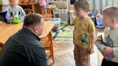 Дорослий чоловік сидить, пригнувшись, біля дітей у дитсадку