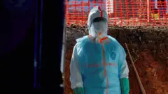 Um profissional de saúde usa equipamento completo de proteção biológica, incluindo macacão impermeável, máscara facial, viseira e luvas, em um ambiente ao ar livre. A figura está posicionada próxima a uma escavação de terra, enquanto uma cerca plástica laranja de isolamento aparece ao fundo. A iluminação destaca o traje de proteção contra o cenário de solo avermelhado e áreas escuras ao redor.