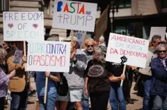 Pessoas protestam na rua contra Donald Trump, portando cartazes em português