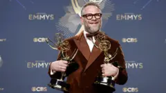 Seth Rogen vestido con un traje burgundy, camisa blanca y pajarita, posa con los cuatro premios Emmy obtenidos por la serie cómica El estudio.