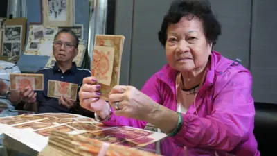 九龍深水埗一公寓樓住客會所內，身穿鮮粉紅色外套的香港索償協會前主席吳溢興遺孀李慧玲（右）舉起一疊紅色的100圓二戰日本軍票（BBC中文圖片）