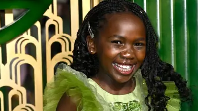 Karis Musongole is stood in front of a green and gold background whilst smiling at the camera. She is wearing a green dress and has long black braided hair