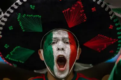 Un hombre con la boca abierta, seguramente está gritando o cantando. Tiene la cara pintada con los colores verde, blanco y rojo, que conforman la bandera de México. Lleva puesto un sombrero de mariachi.