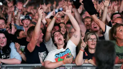 Fãs do Oasis cantam durante show com os braços para cima. 