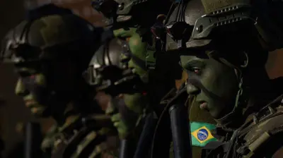 Close de quatro militares brasileiros usando capacetes, equipamentos táticos e pintura de camuflagem no rosto. Eles aparecem perfilados lado a lado, em posição de atenção, com a bandeira do Brasil visível no uniforme de um deles e iluminação que destaca seus rostos.