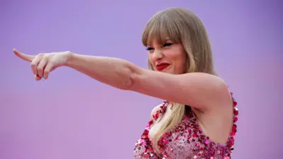 Taylor Swift wskazuje w dal z szerokim uśmiechem, mrużąc oczy spod blond grzywki. Ma na sobie dopasowany, błyszczący różowy gorset oraz czerwoną szminkę. Tło w różowo-fioletowym gradiencie współgra z jej stylizacją