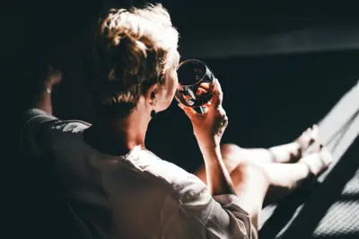 Mulher bebendo taça de vinho tinto, vestindo um roupão de seda e brincos