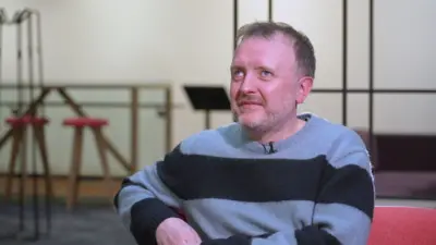 Chris McCausland wearing a stripey jumper and smiling sits on a sofa in a BBC office.