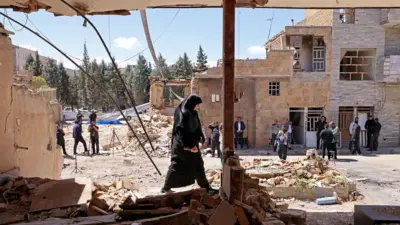 A woman walks past the residential buildings that were damaged by recent strikes at Vahdat town in Karaj, southwest of Tehran on 3 April, with bystanders and other buildings in the background against a sunny blue sky.
