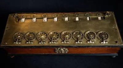 Máquina construída em madeira e bronze, com rodas numeradas que permitem os cálculos