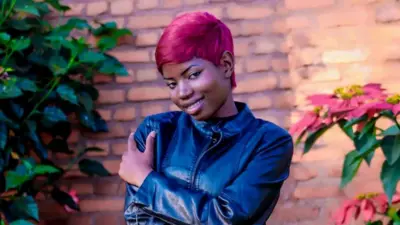 Umuririmvyikazi Marlène Kamikazi batazira “B Charly” yambaye ikote ry'umukara, inyuma ye hari inzu y'amatafari, iruhande rwe hari amashugwe. 