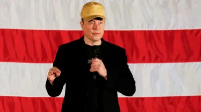 Elon Musk, wearing a gold MAGA hat, speaks into a microphone while standing in front of an American flag.