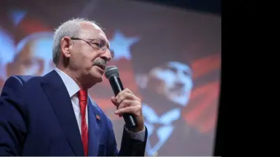Kemal Kılıçdaroğlu elinde mikrofonla ayakta konuşuyor. Kırmızı bir kravat takmış arkasında fonda kalpaklı bir Atatürk fotoğrafı var