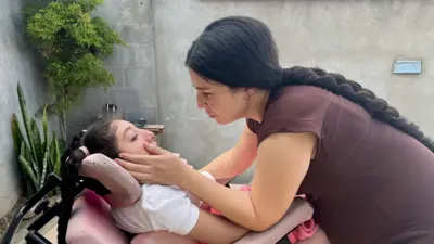 Uma mulher olhando para a filha que está em uma cadeira de rodas. Ela segura seu rosto.