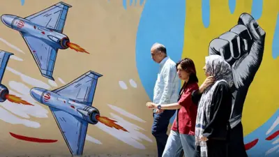 Homem e mulher idosos e mulher mais jovem andam em frente a um mural em Teerã com vários drones e um pulso erguido pintados