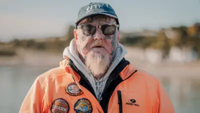 Angler Jay Lewis who is wearing a grey cap, orange waterproof jacket, sunglasses and has facial tattoos and a nose ring