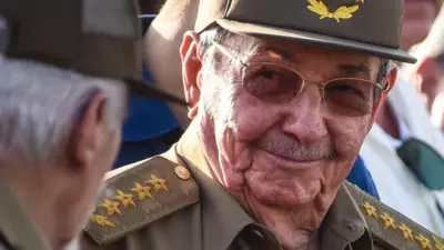 Primer plano de Raúl Castro, con gafas de sol y uniforme, sonriendo a otra persona