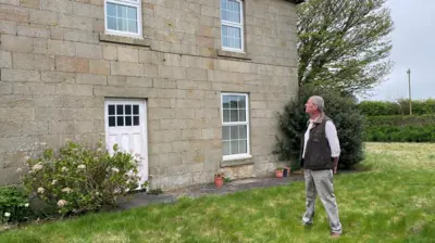 Martin Beal is looking at his house which is a Georgian stone built farmhouse with a white door and windows. He is a man, about 60 years of age, wearing a waist coat, white shirt and light coloured trousers.