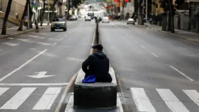 Каракас, 5 января 2026 года: на барьере, разделяющем проезжую часть улицы, прямо рядом с пешеходным переходом сидит мужчина в бейсболке. Машин на улице немного, людей тоже