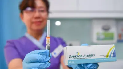 A healthcare worker in purple uniform and blue medical gloves holds a syringe toward the camera while holding a white vaccine box in the other hand 