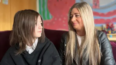 Hannah and Ashly are sitting in a classroom looking at each other. Ashly, the mother, has long blonde hair and is wearing a grey leather jacket and white blouse. She is smiling at her daughter who is looking into the distance. Hannah has shoulder-length dark hair and is wearing a white school shirt without a tie and black hoodie. 