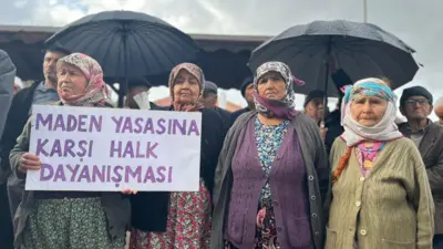 Dört kadın, geleneksel köy kıyafetleriyle yanyana duruyor, en soldakinin elinde Maden Yasasına Karşı Halk Dayanışması yazılı bir pankart var. Geri iki siyah şemsiye ve diğer köylüler flu. 